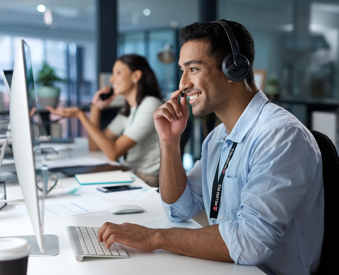 Photo of a man wearing a headset looking at a computer screen, and a woman in the background speaking on a desk phone