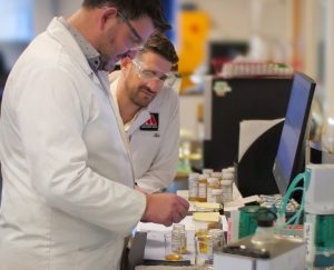 Photo of 2 men in the lab wearing white lab coats and safety glasses, looking at some small bottles of oil in front of them, and a computer monitor