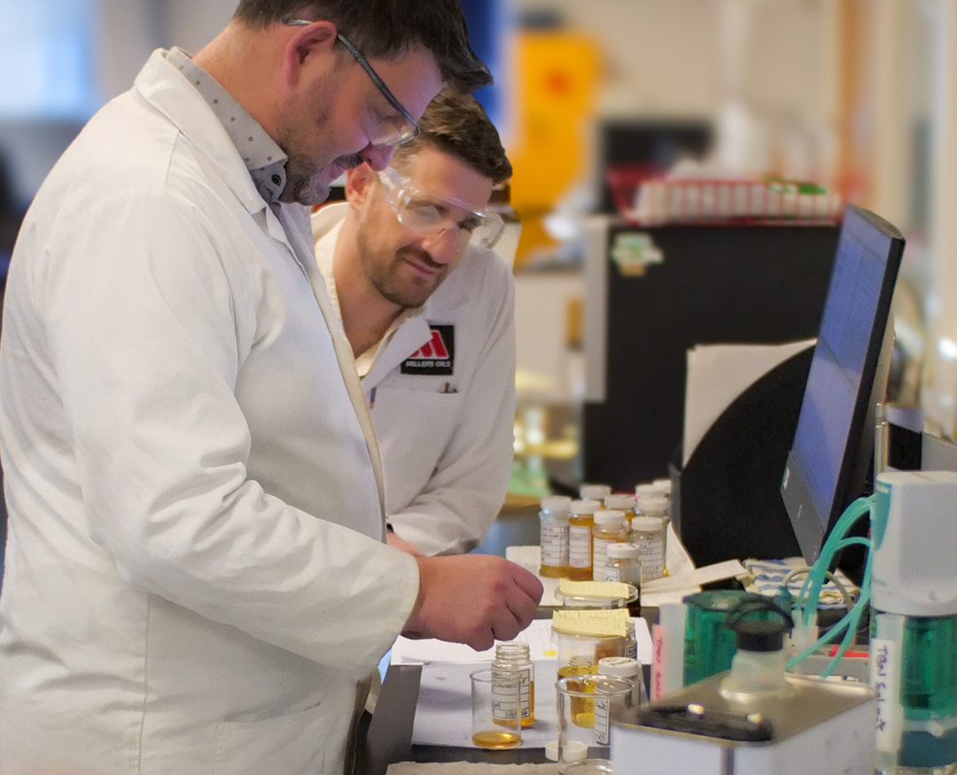 Photo of 2 men in the lab wearing white lab coats and safety glasses, looking at some small bottles of oil in front of them, and a computer monitor